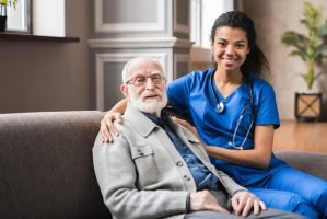 Front view portrait of caring afro nurse taking care of an elderly caucasian man grandfather grandpa indoors. Happy female doctor consulting old man in clinic.